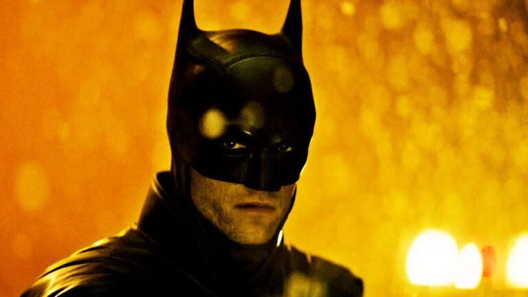 Robert Pattinson als Batman in schwarzer Maske und Rüstung vor einem intensiv orange leuchtenden Hintergrund. Sein Gesicht liegt teilweise im Schatten, während helle Lichtreflexe hinter ihm sichtbar sind.