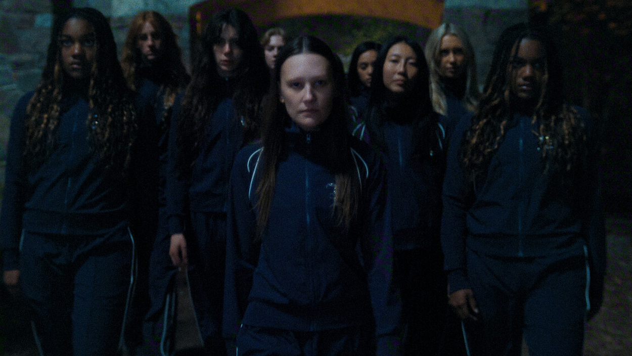 Eine Gruppe junger Frauen in dunklen Trainingsanzügen steht nachts dicht zusammen und blickt ernst nach vorn.