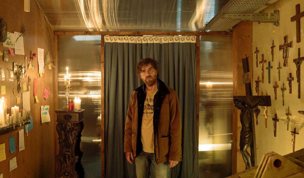 Filmszene aus The Holy Boy: Ein bärtiger Mann in brauner Jacke steht in einem schmalen Raum vor einem dunklen Vorhang. An den Wänden hängen zahlreiche Kruzifixe, Heiligenbilder und handgeschriebene Zettel. Brennende Kerzen tauchen den Raum in warmes, gelbliches Licht.