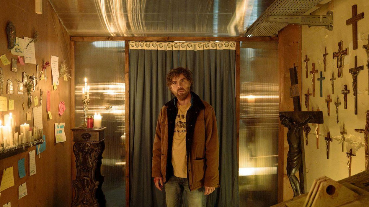 Filmszene aus The Holy Boy: Ein bärtiger Mann in brauner Jacke steht in einem schmalen Raum vor einem dunklen Vorhang. An den Wänden hängen zahlreiche Kruzifixe, Heiligenbilder und handgeschriebene Zettel. Brennende Kerzen tauchen den Raum in warmes, gelbliches Licht.