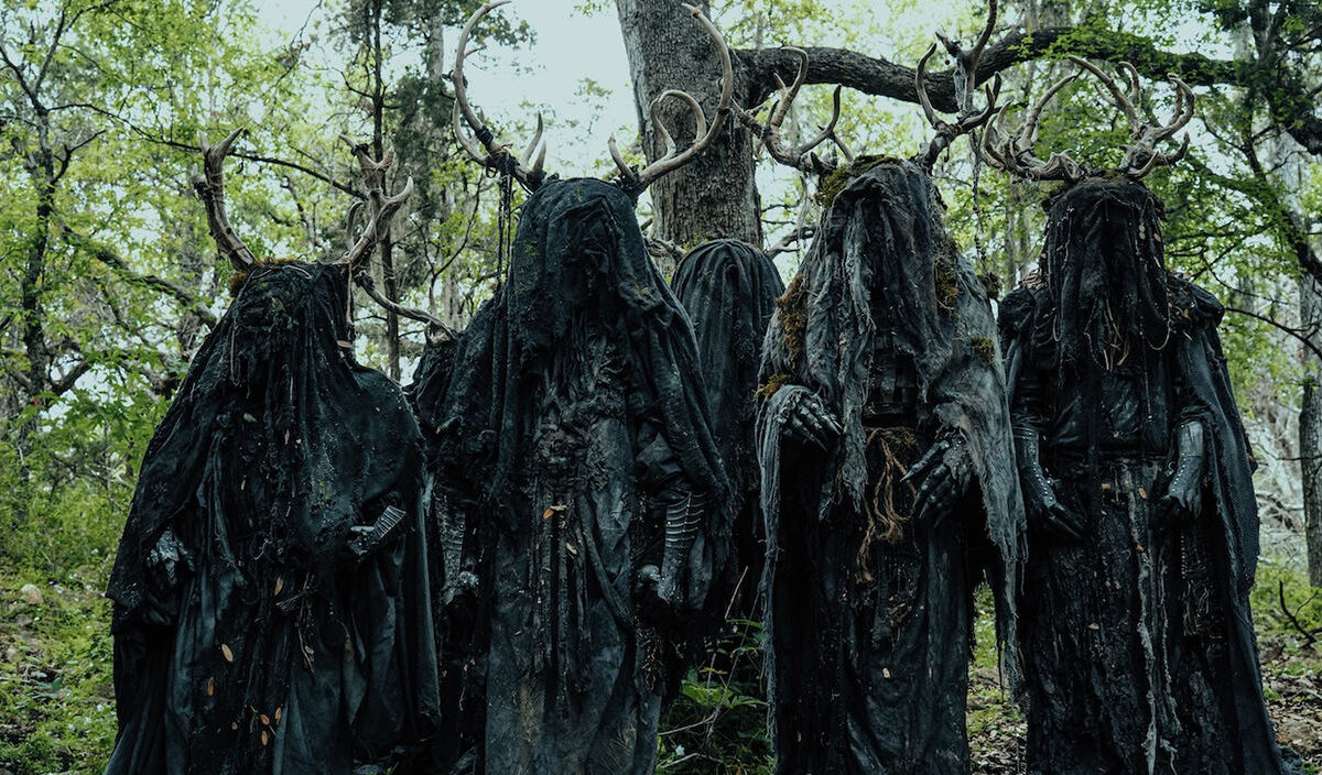 Mehrere verhüllte dunkle Gestalten mit großen Geweihen stehen in einem Wald. Die Figuren tragen schwarze, zerlumpte Gewänder und wirken wie unheimliche Kultwesen.