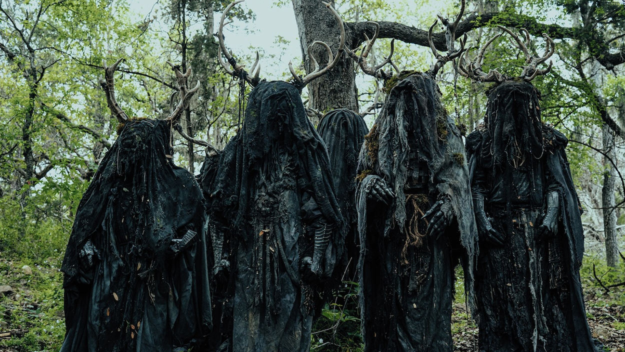 Mehrere verhüllte dunkle Gestalten mit großen Geweihen stehen in einem Wald. Die Figuren tragen schwarze, zerlumpte Gewänder und wirken wie unheimliche Kultwesen.