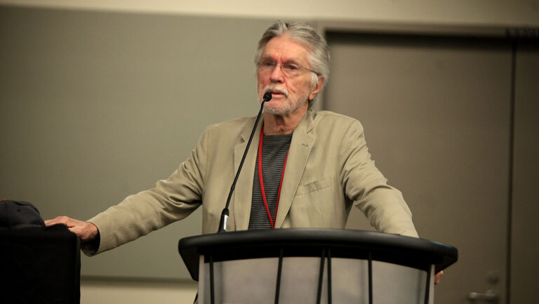 Tom Skerritt mit grauem Haar, Bart und beigem Sakko spricht bei einer Veranstaltung im Dezember 2014 hinter einem Rednerpult in ein Mikrofon.
