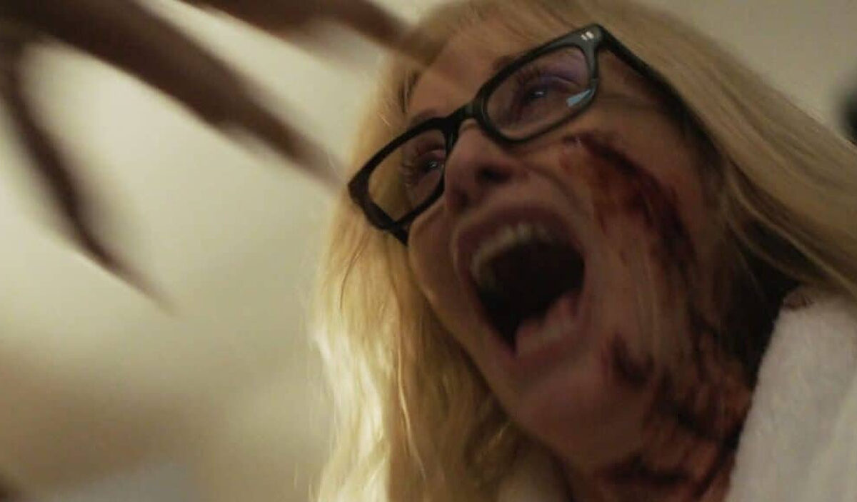 Horrorszene aus Trauma Or, Monsters All: Eine blonde Frau mit schwarzer Brille schreit mit weit geöffnetem Mund. Blutige Spuren ziehen sich über ihre rechte Wange und ihren Hals, während im unscharfen Hintergrund eine bedrohliche, klauenartige Form zu erkennen ist.