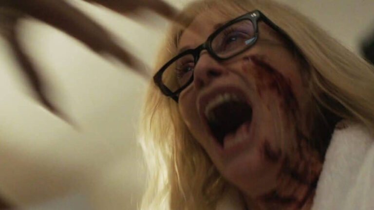 Horrorszene aus Trauma Or, Monsters All: Eine blonde Frau mit schwarzer Brille schreit mit weit geöffnetem Mund. Blutige Spuren ziehen sich über ihre rechte Wange und ihren Hals, während im unscharfen Hintergrund eine bedrohliche, klauenartige Form zu erkennen ist.