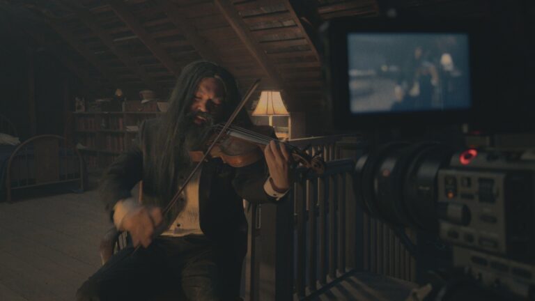 Ein langhaariger Musiker spielt auf einem Dachboden Geige, während ihn eine Videokamera in V/H/S/MIXTAPE filmt.