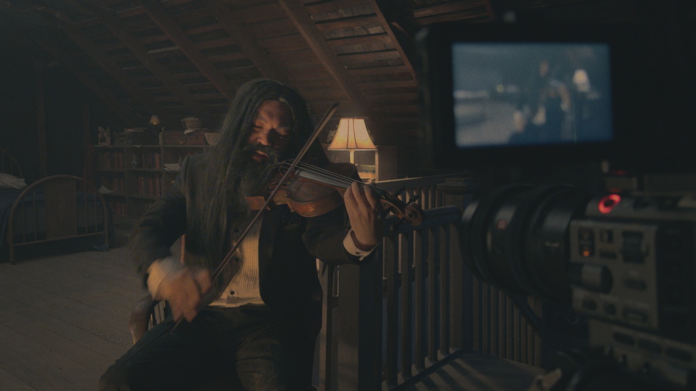 Ein langhaariger Musiker spielt auf einem Dachboden Geige, während ihn eine Videokamera in V/H/S/MIXTAPE filmt.