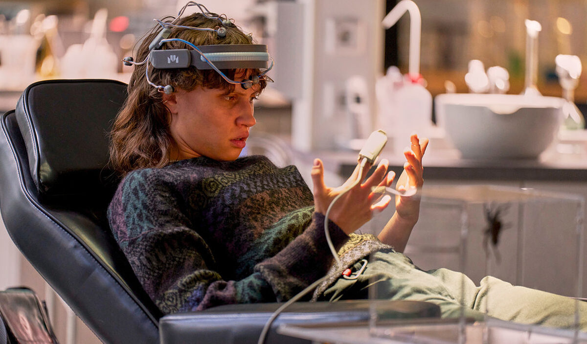 Luna Jordan als Emilia in der ZDF-Science-Fiction-Serie Wake Up: Mit einem technischen Gerät am Kopf sitzt sie in einem Behandlungsstuhl und betrachtet verwundert ihre Hände, während Hevis Bewusstsein ihren Körper kontrolliert.