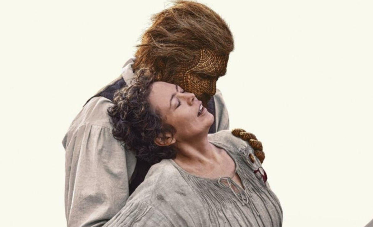 Filmszene aus Wicker: Eine lachende Frau mit dunklem, hochgestecktem Haar und grauem Kleid lehnt sich zurück an einen großen Mann mit geflochtenem Gesicht, braunem Haar und heller Kleidung. Der Weidenmann neigt seinen Kopf zu ihr.