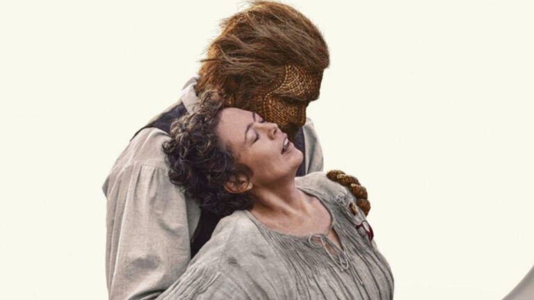 Filmszene aus Wicker: Eine lachende Frau mit dunklem, hochgestecktem Haar und grauem Kleid lehnt sich zurück an einen großen Mann mit geflochtenem Gesicht, braunem Haar und heller Kleidung. Der Weidenmann neigt seinen Kopf zu ihr.