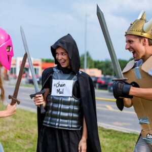 Drei junge Erwachsene in selbstgebastelten LARP-Kostümen aus Pappe, Plastik und Humor liefern sich ein episches Straßenduell. Kartonrüstung trifft auf pinken Helm und ein „@chain mail“-Shirt – ein brillanter Mix aus Fantasy-Parodie, Wortwitz und Alltagswahnsinn. Perfekt für Meme-Galerien mit Herz und Hirn.