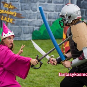 Improvisiertes Fantasy-Duell zwischen einer pink gekleideten Prinzessin mit Spielzeugkrone und einem Ritter mit Papp-Rüstung und Footballhelm; beide kämpfen mit Spielzeugschwertern auf einer Wiese vor einer bemalten Kulisse mit der Aufschrift „DJ JJ’s Dragon’s Lair“.